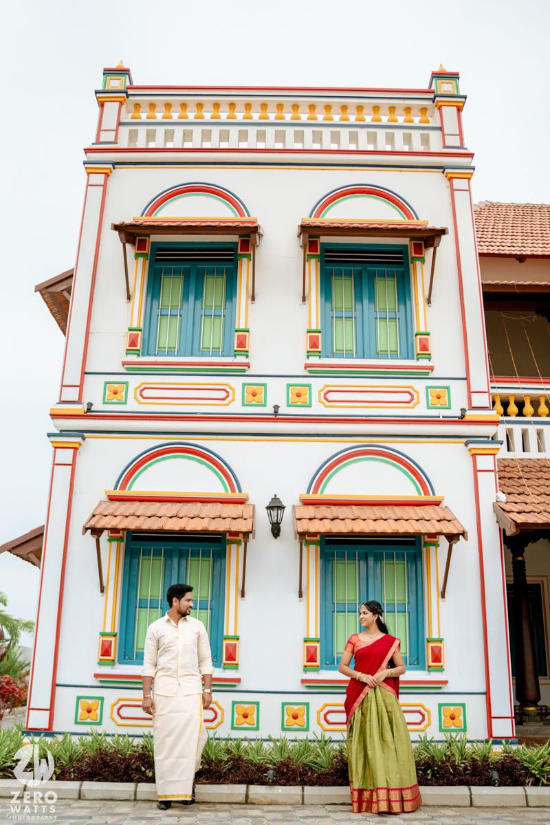 South Indian couple in traditional attire standing before a colorful heritage building facade.