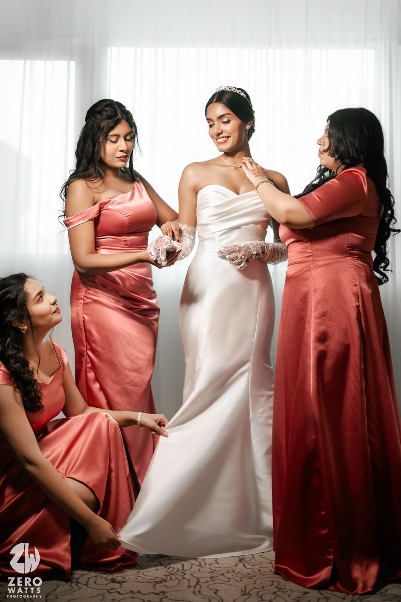 Bride in white gown gets ready with bridesmaids in coral satin dresses helping with accessories.