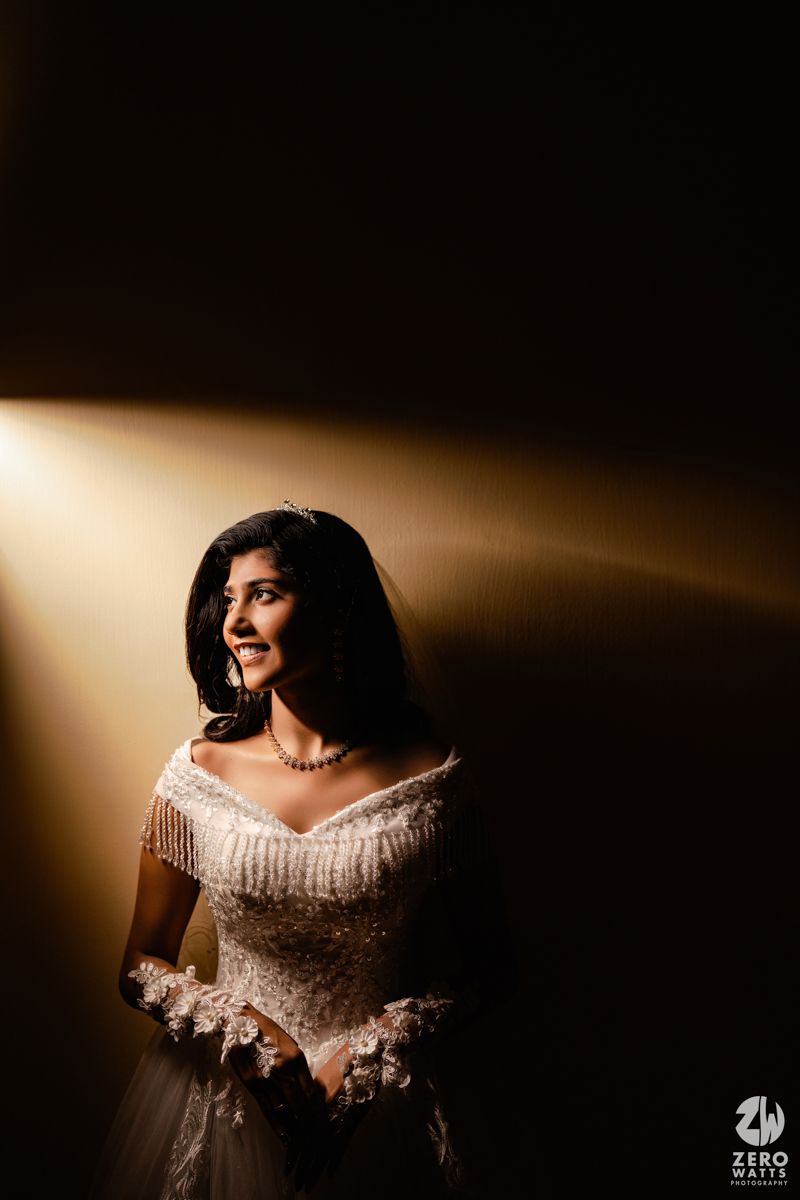 Bride in white off-shoulder gown with tiara smiles under dramatic golden spotlight against dark background.