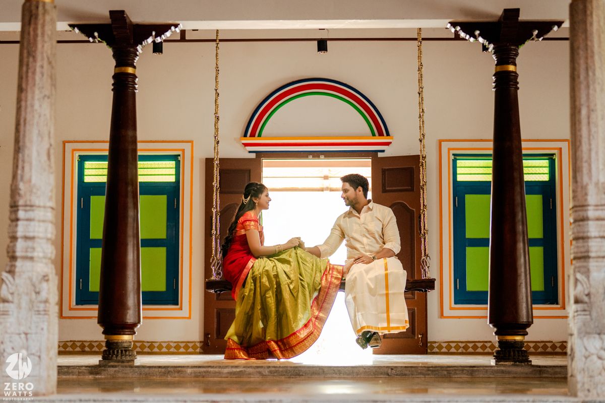Couple in traditional South Indian attire sitting on a wooden swing in a colorful heritage home doorway.