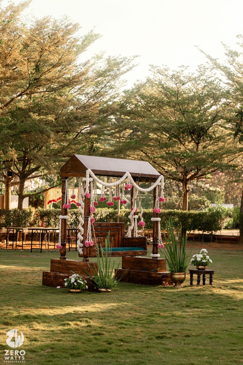 Rustic wooden mandap decorated with pink flowers and white garlands in a lush green outdoor garden wedding venue.