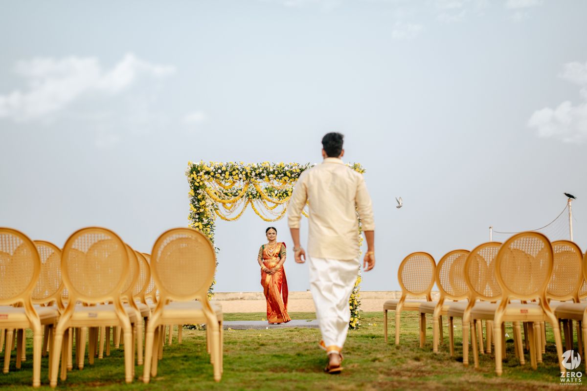 Indian bride and groom walk toward a floral mandap surrounded by rattan chairs at an outdoor wedding ceremony.