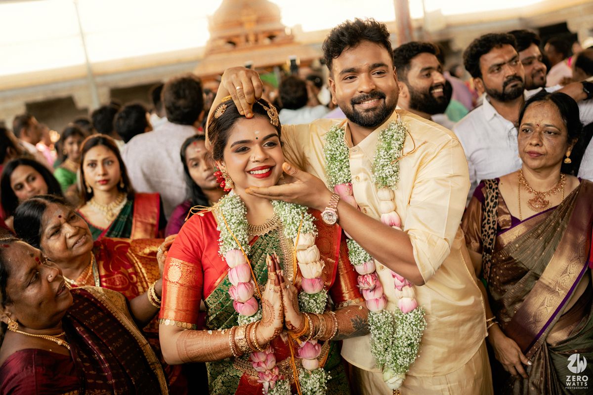 South Indian wedding couple in traditional attire exchanging garlands surrounded by joyful family members at outdoor ceremony.