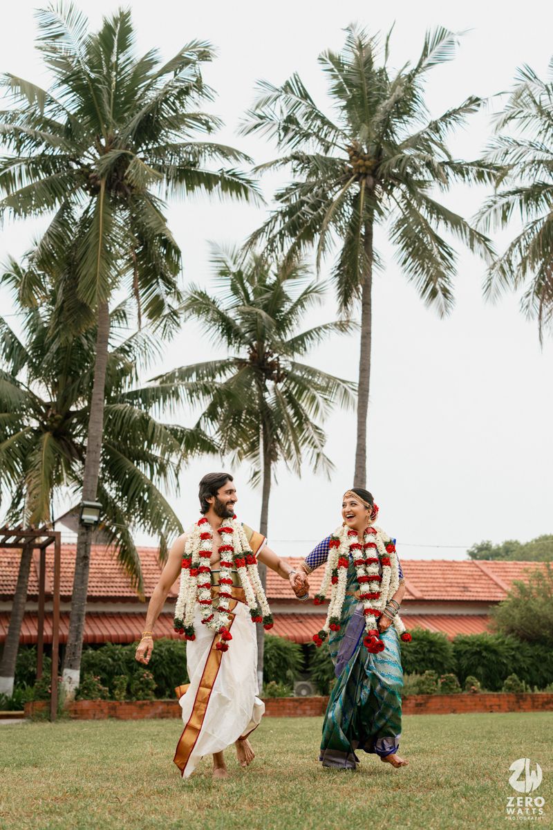 Indian couple in traditional wedding attire with flower garlands walking hand in hand on grass with palm trees backdrop.