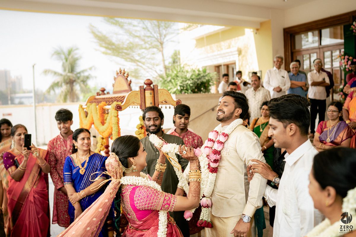 Colorful South Indian wedding ceremony with guests in traditional attire, groom in cream sherwani exchanging garlands outdoors.