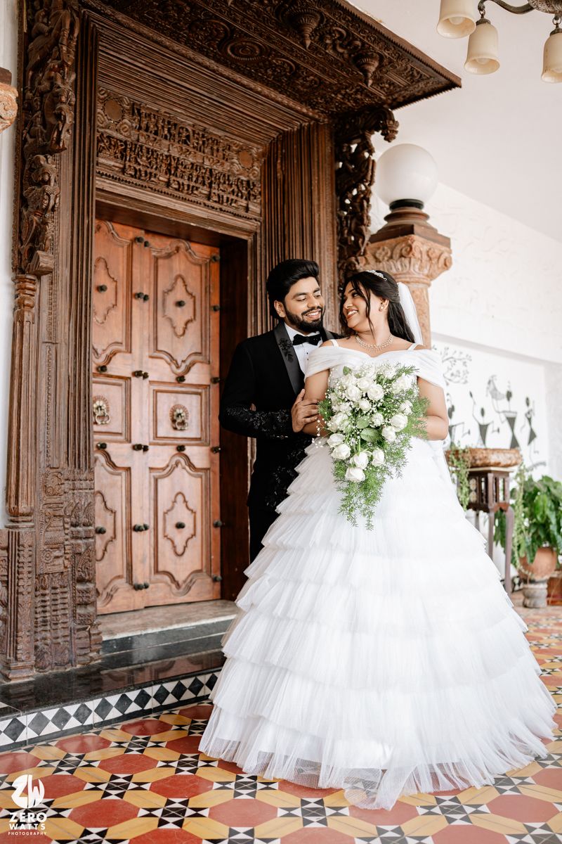 Bride in white ruffled gown holds bouquet beside groom in tuxedo at ornate wooden door.