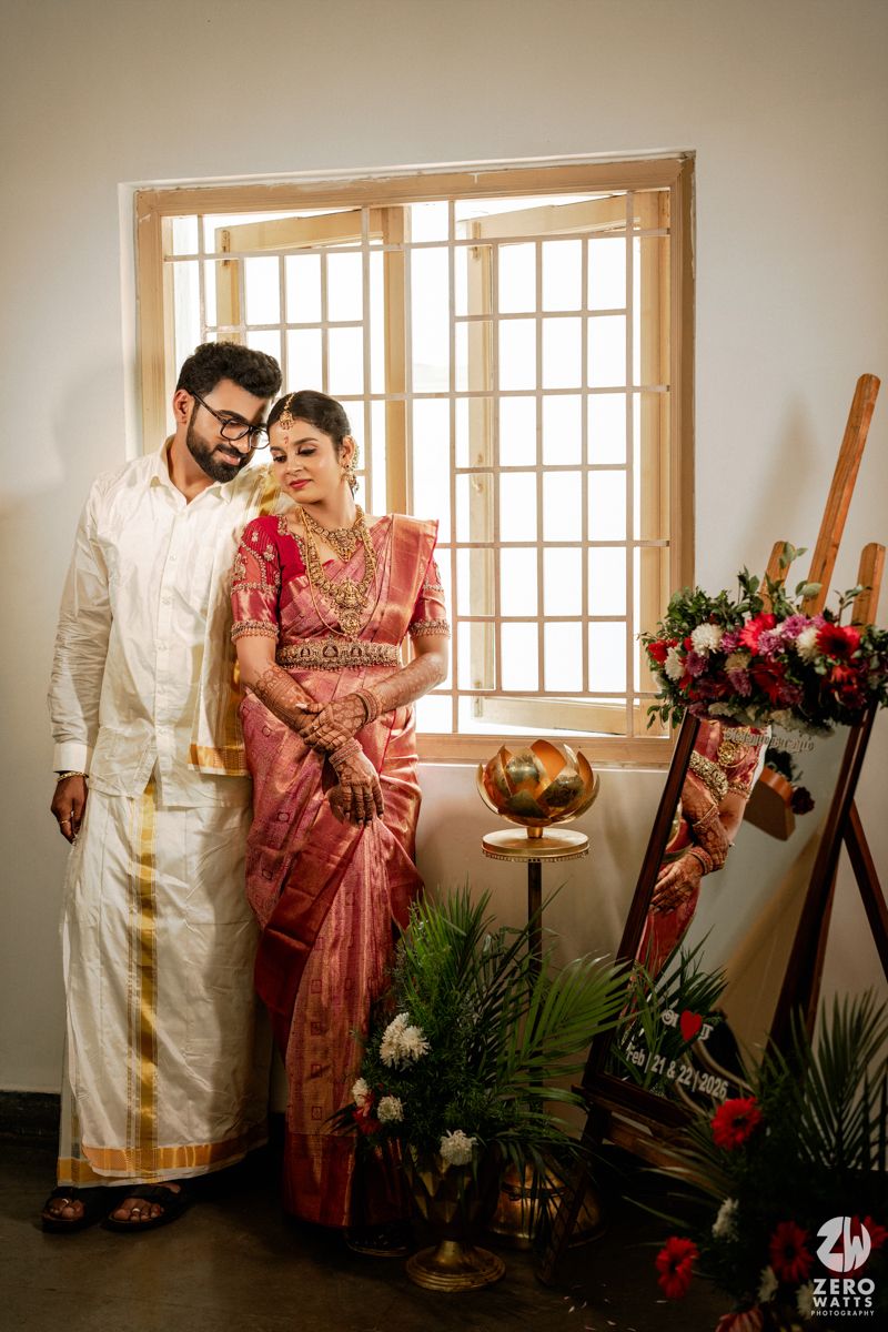 South Indian wedding couple posing by window, groom in white dhoti and bride in pink Kanjivaram saree.