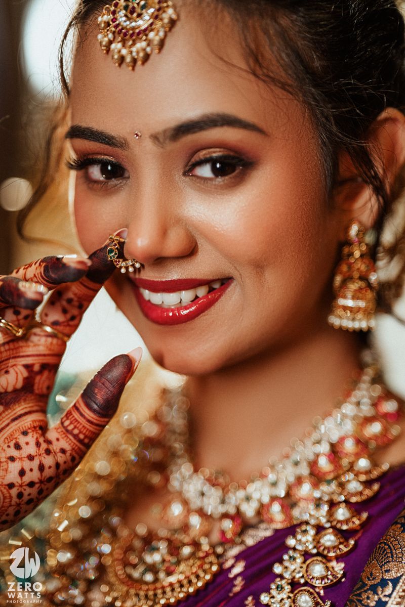 Smiling Indian bride in gold jewelry adjusts nose ring, wearing mehndi and traditional bridal makeup.