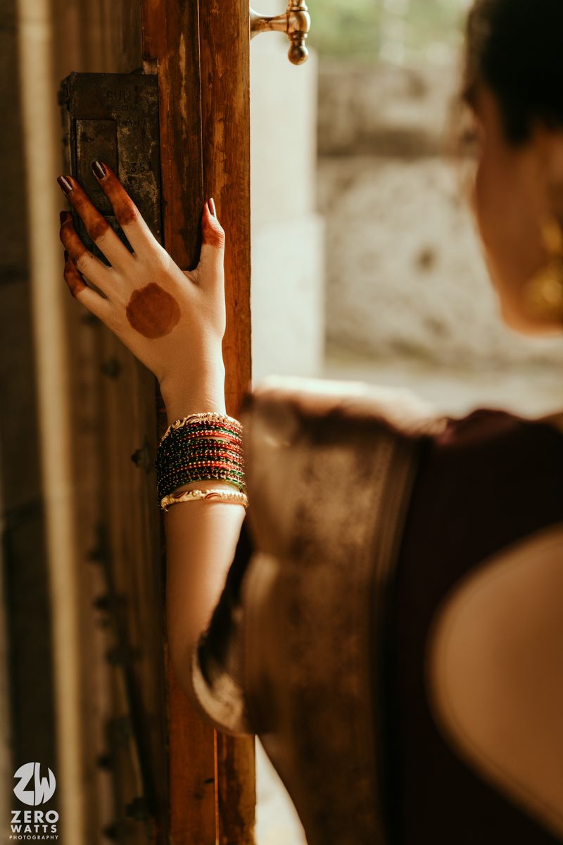 A woman's hand adorned with henna, bangles, and long nails rests on a rustic wooden door in warm light.