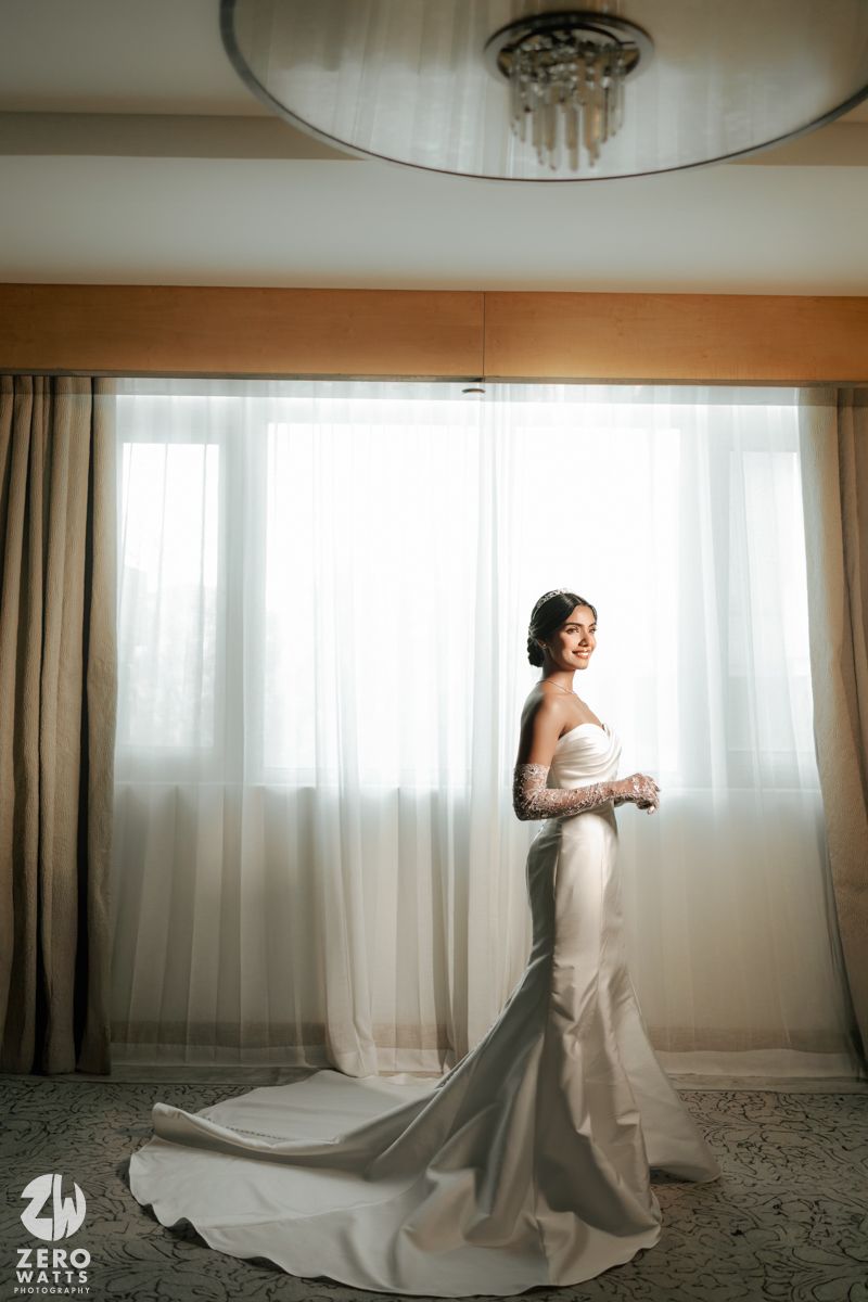 Bride in white mermaid wedding gown with lace sleeves standing by sheer curtained window.