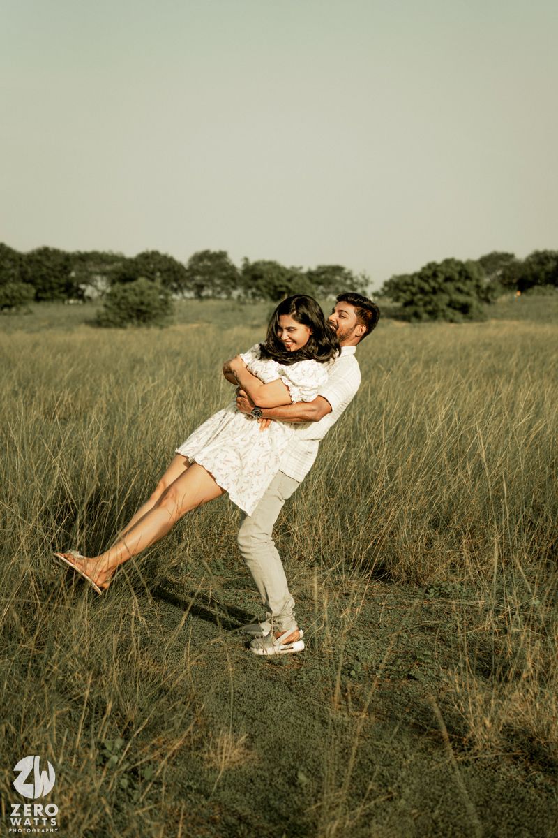 Playful couple in white clothing embracing and spinning in a grassy meadow at sunset.