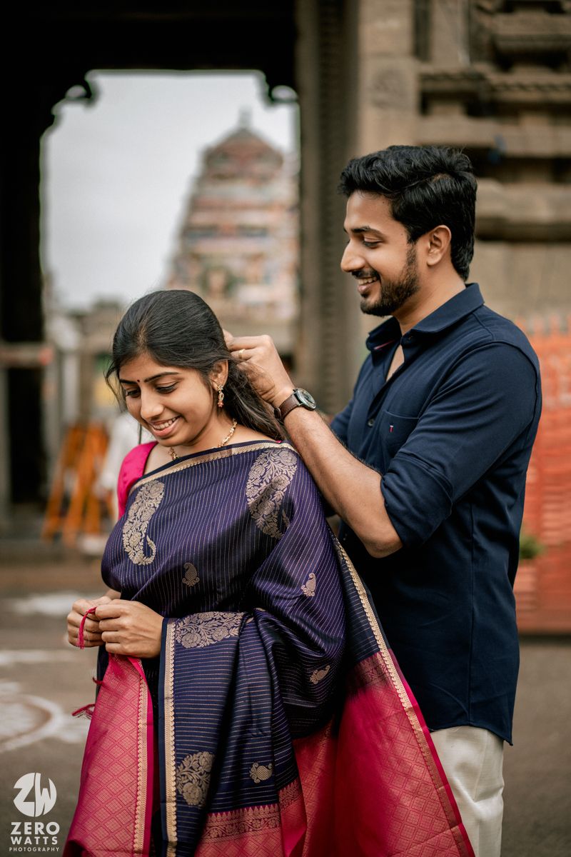 A couple in traditional Indian attire share a tender moment as he helps adjust her sari in an outdoor setting.