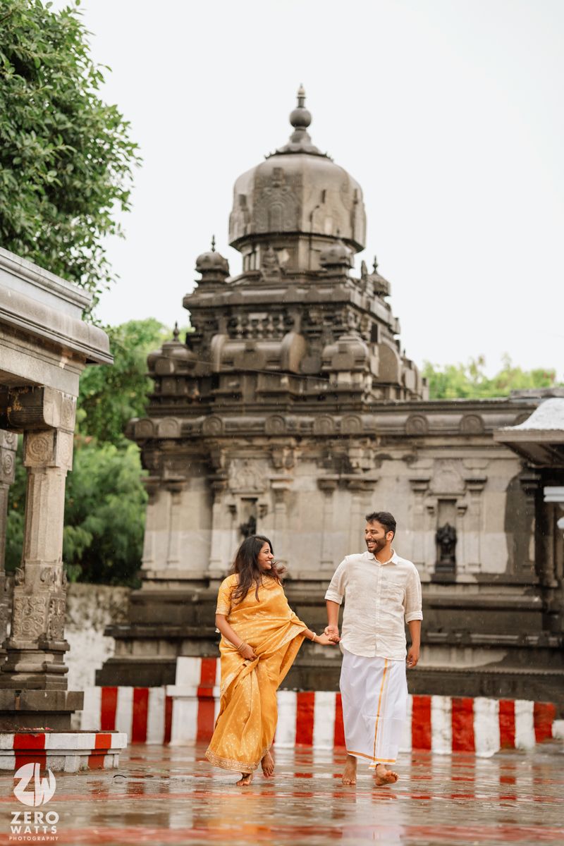 A couple in white and orange outfits pose together at a historic stone temple with domed architecture.