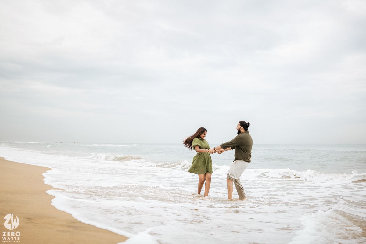 A couple dances playfully together on a cloudy beach with waves crashing in the background.