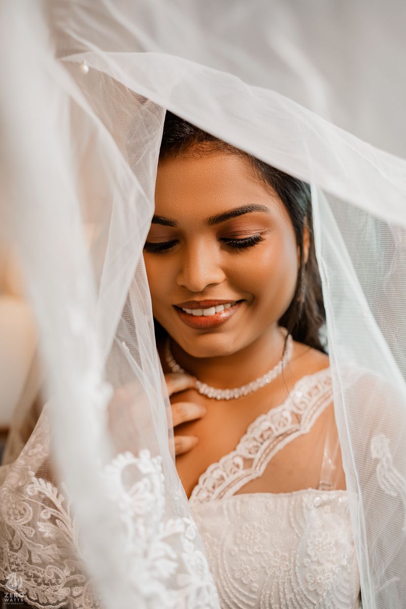 A bride in a white lace wedding dress and veil smiles softly while wearing pearl jewelry.