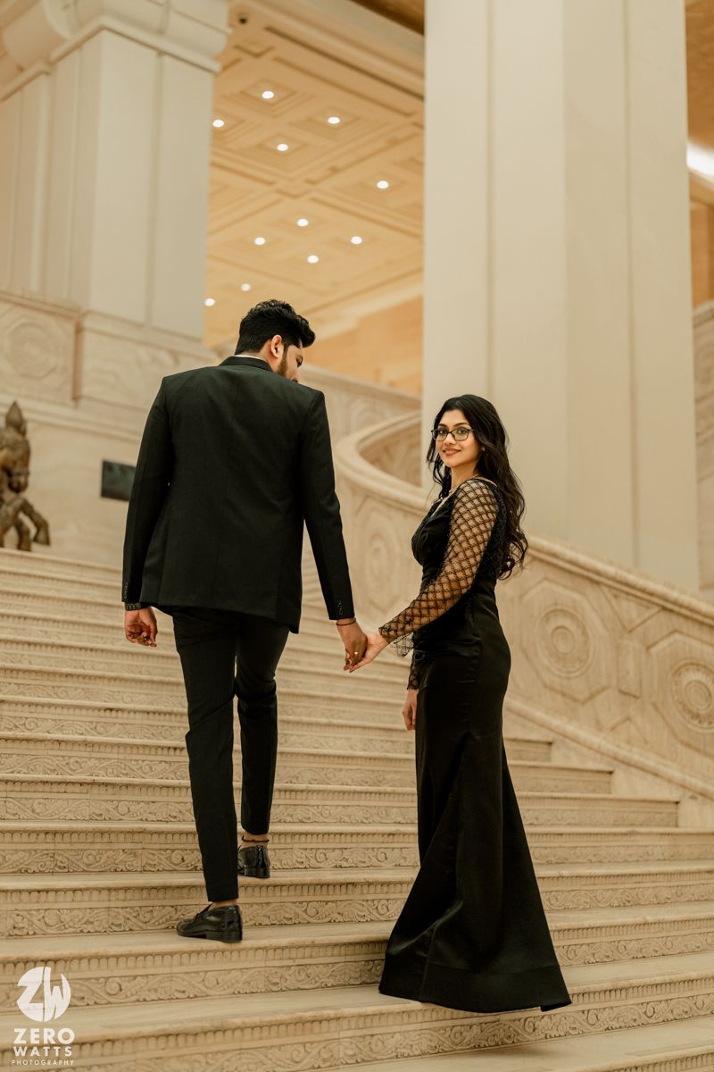 A couple in formal attire ascends a grand marble staircase in an elegant building with ornate architectural details.