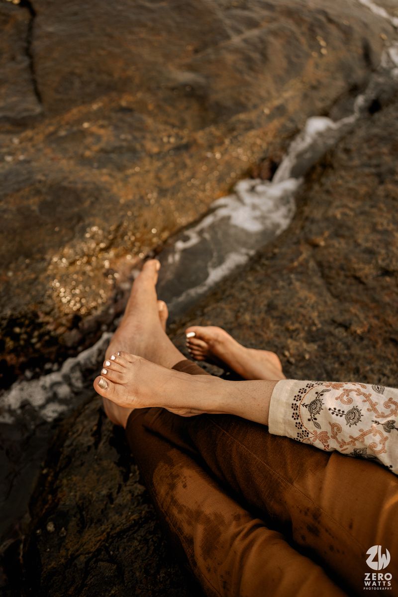 Close-up of bare feet with lace dress detail resting on textured brown rocks near water.