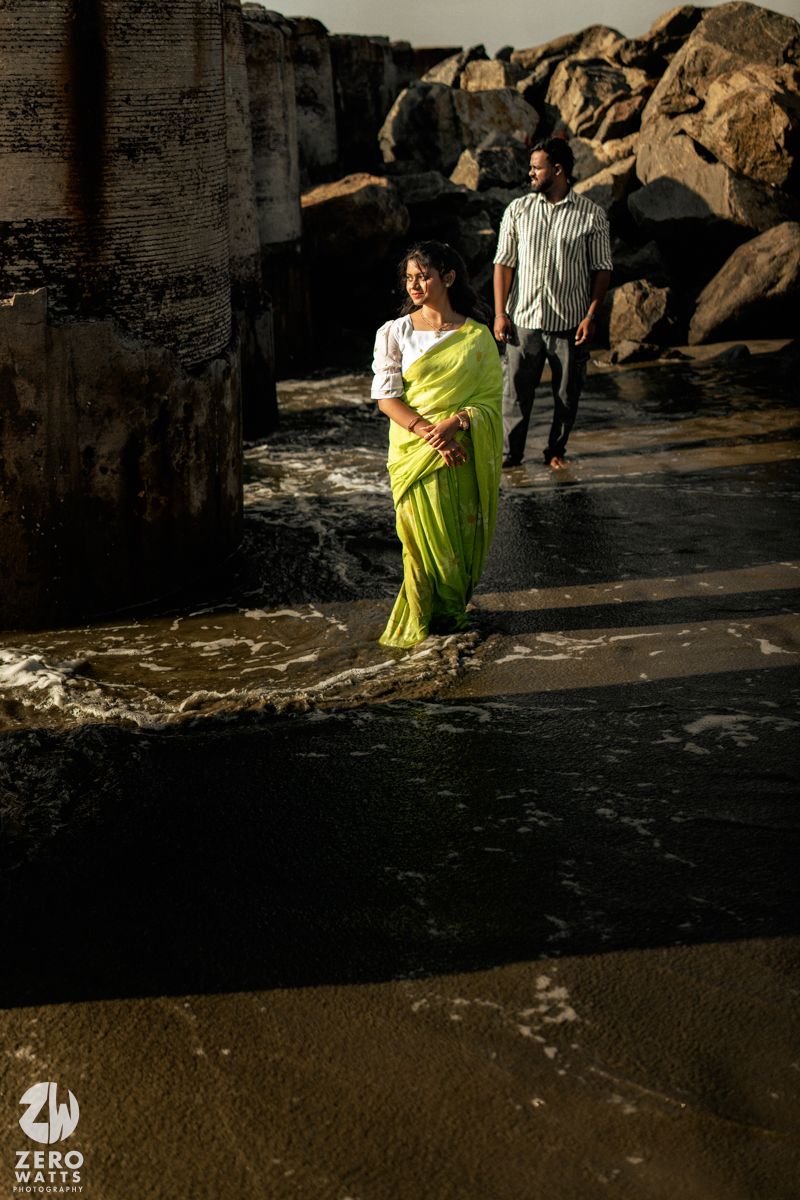 Couple in traditional attire poses near ocean waves and stone structures.