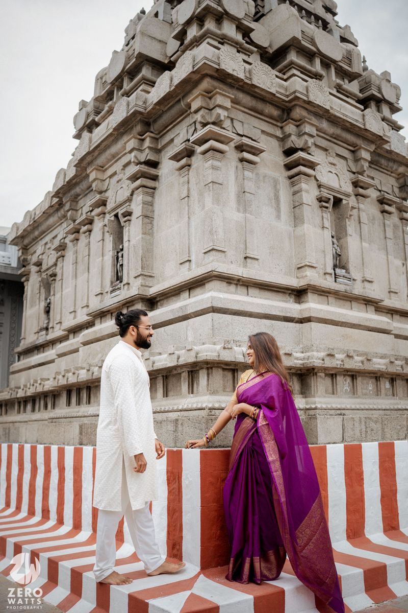 A couple in traditional Indian attire stands in front of an ornate temple with red and white striped walls.