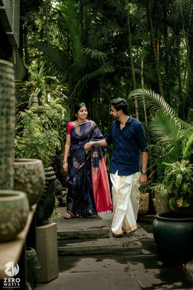 A couple walks through a lush tropical garden pathway lined with palm plants and decorative pots.