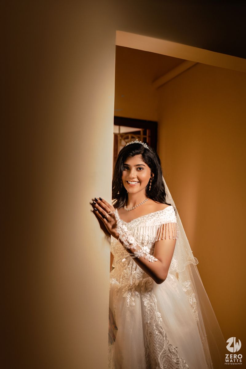 Radiant bride in white lace gown and tiara smiles joyfully, peeking around a golden wall on her wedding day.