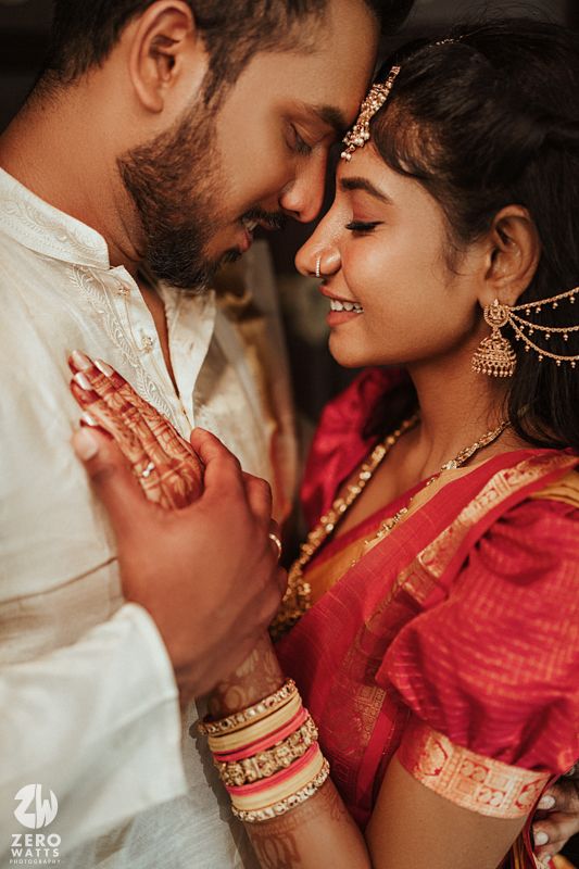 South Indian bride in red silk saree with gold jewelry shares an intimate moment with groom in white kurta.