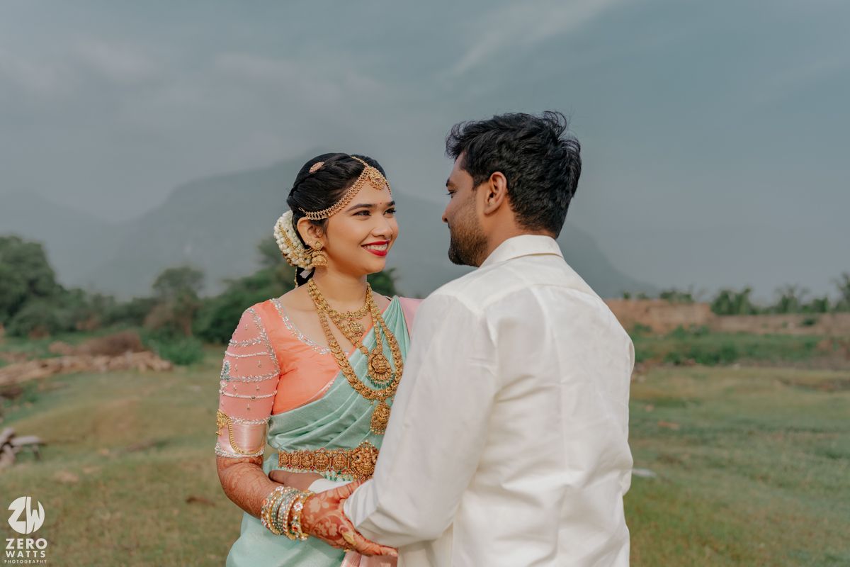 Indian bride in green saree and groom in white kurta share a romantic moment outdoors with mountains in background.