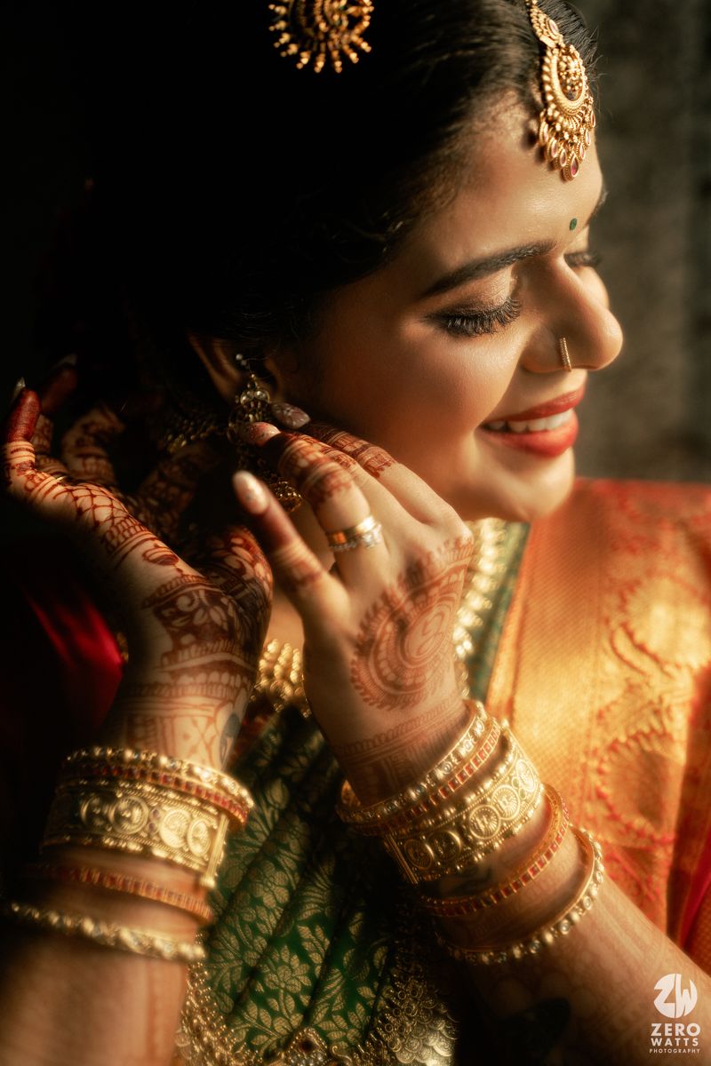 South Indian bride adorned in gold jewelry and mehndi, smiling while adjusting her earring in a red and green silk saree.