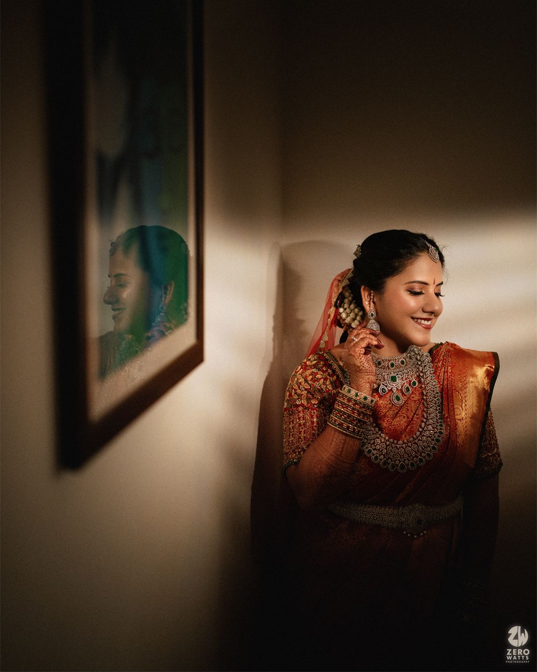 Smiling bride in an orange silk saree and ornate jewelry poses beside a framed picture, lit by soft window light.