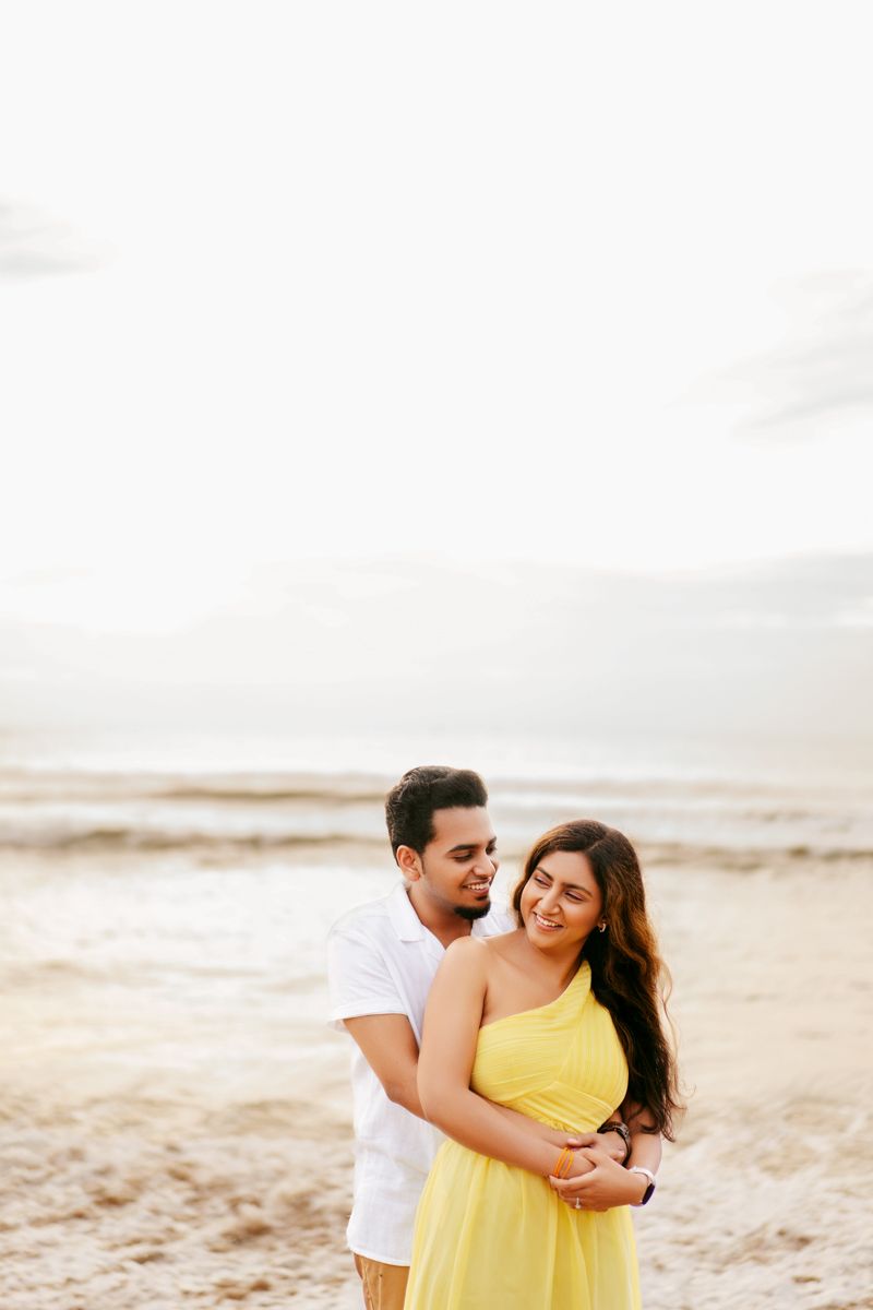 Romantic couple embraces on sandy beach at sunset, with person in yellow dress and white shirt.