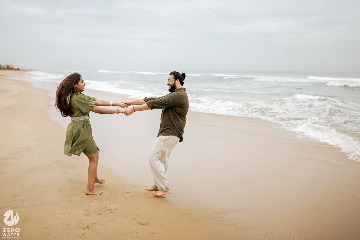 Two people dance playfully on a sandy beach with ocean waves in the background.