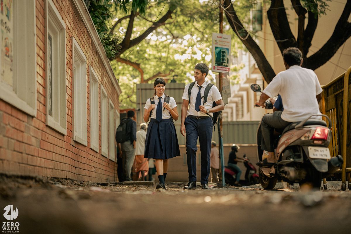 Students in retro uniforms chat on a brick-lined pathway under trees.