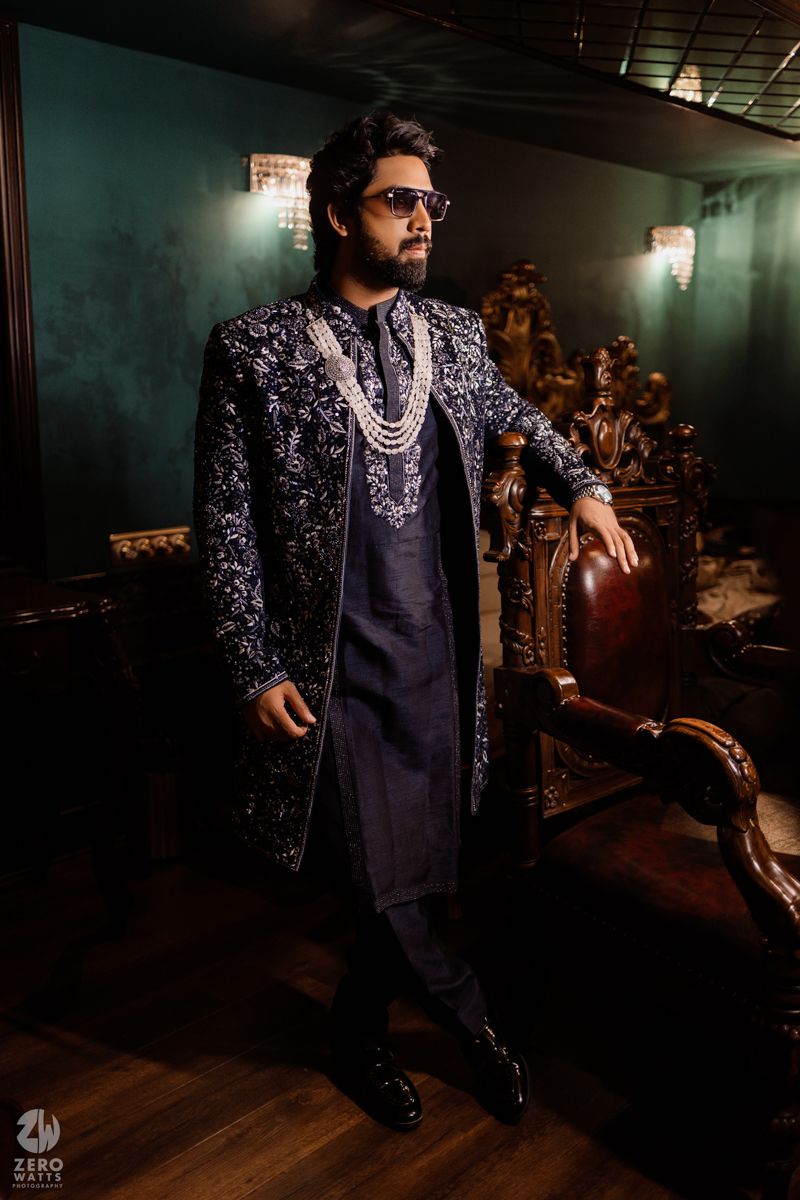Groom in dark embroidered sherwani and sunglasses poses dramatically in moody lighting.