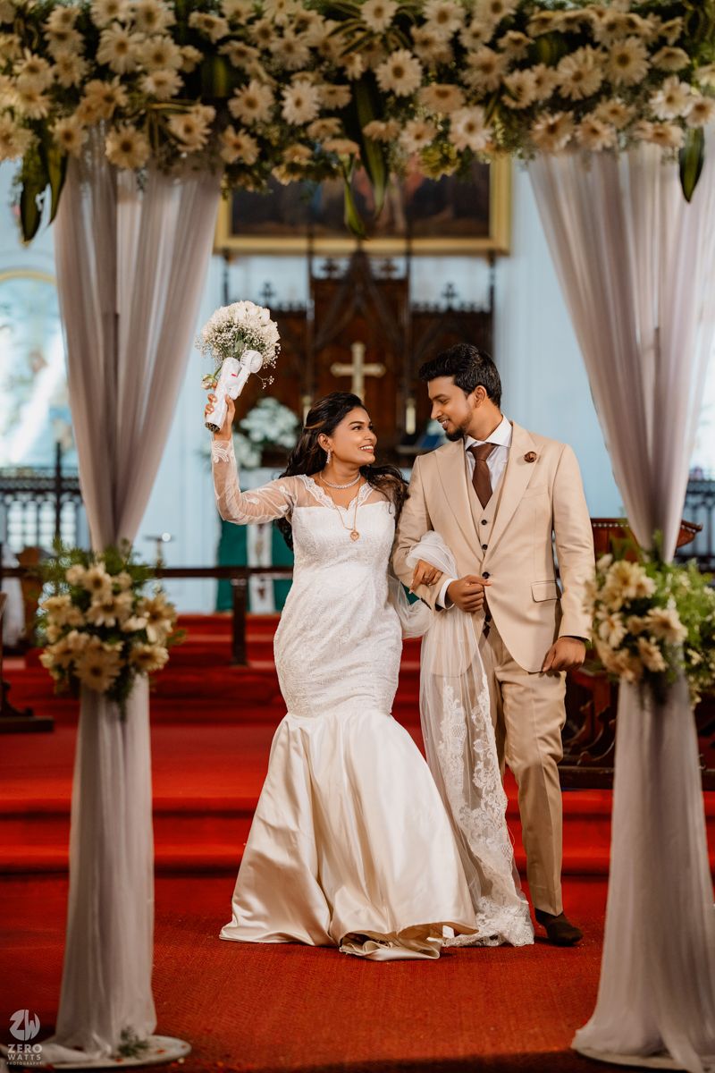 A couple celebrates during their wedding ceremony under a floral arch with white drapes and red carpet in a church setting.