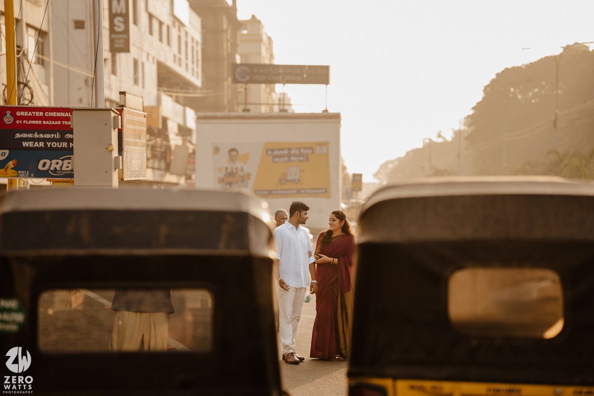Two people stand between auto rickshaws on a sunlit street in India during golden hour.