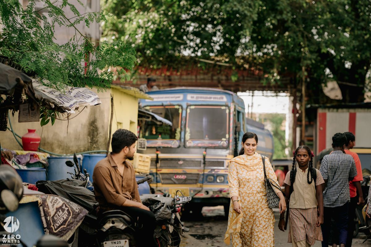A street scene in India with a woman in gold dress walking past parked vehicles and pedestrians.
