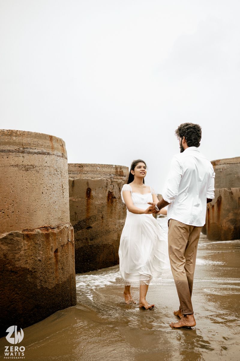 Couple in white outfits holding hands barefoot on a wet rocky beach shore, romantic pre-wedding photoshoot.