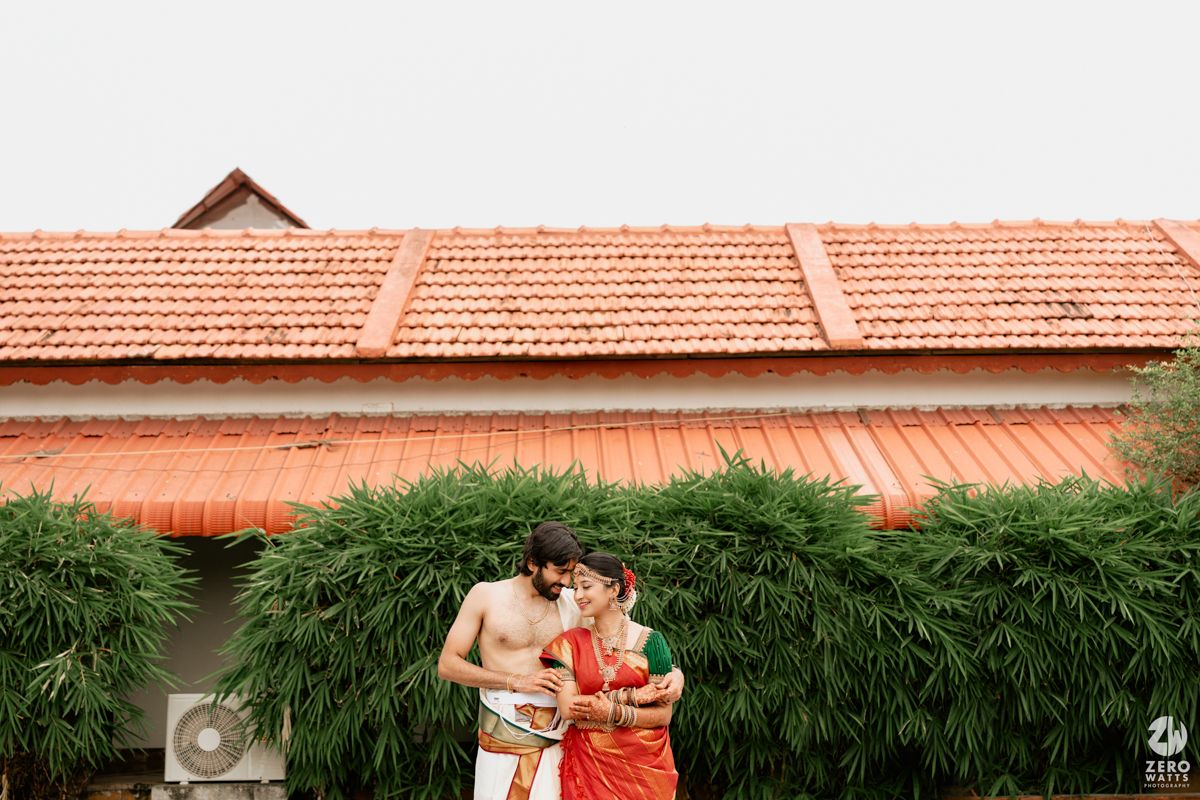 South Indian bride in red saree and groom share romantic moment outdoors with tiled roof background.