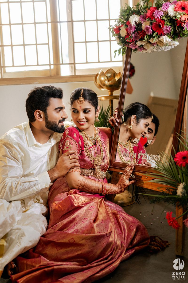 South Indian wedding couple sitting by ornate mirror with floral decor, bride in red silk saree.