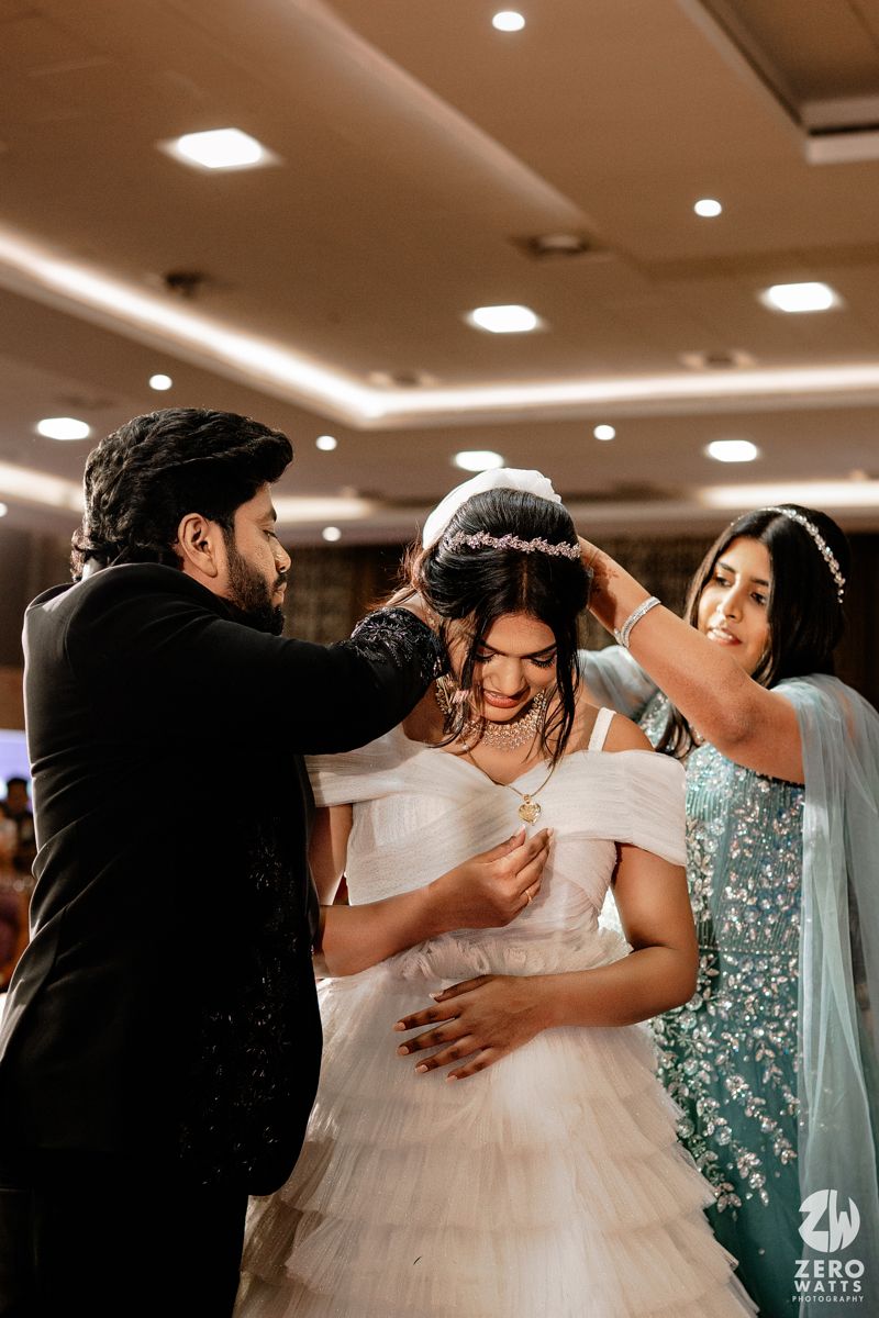 Bride in white gown gets floral crown placed by man in black suit while woman in teal saree assists at wedding.