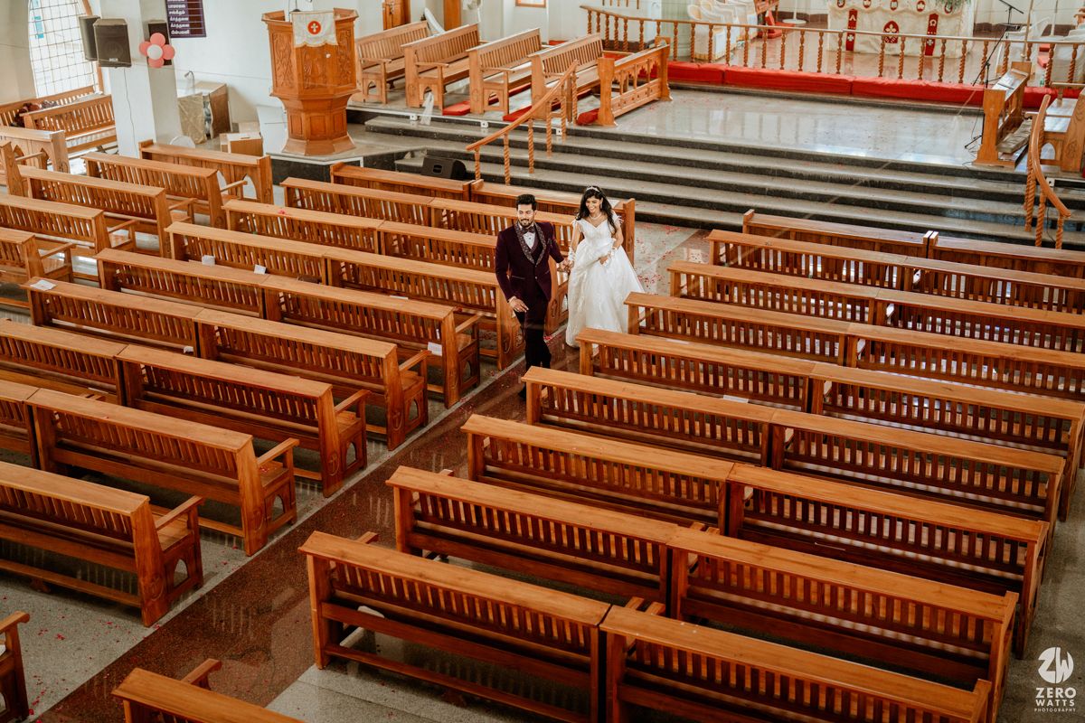 Top-down view of wedding couple strolling through sunlit church nave lined with polished wooden benches.