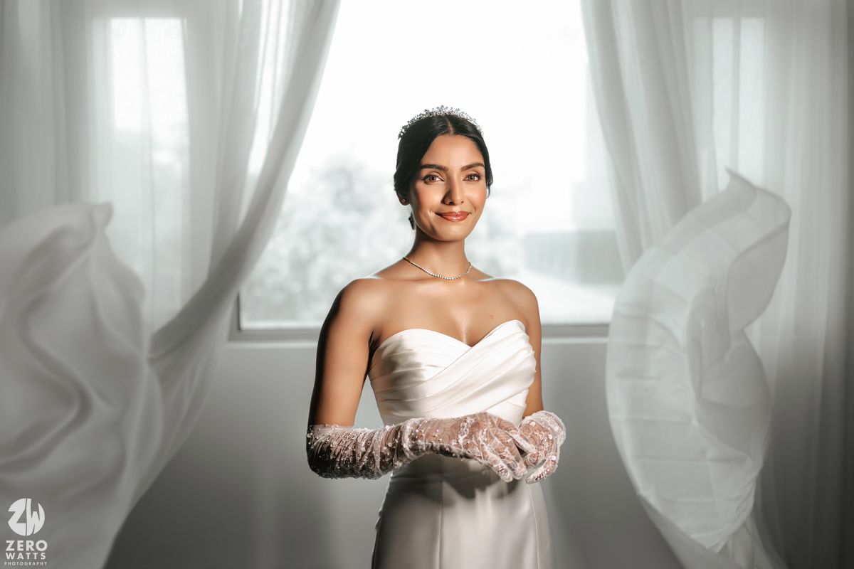 Bride in white strapless gown and lace gloves smiling by billowing curtains.