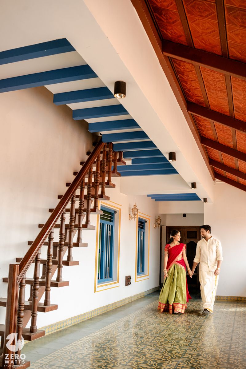 Indian couple in traditional attire holding hands near a wooden staircase with blue accents and tiled floor.