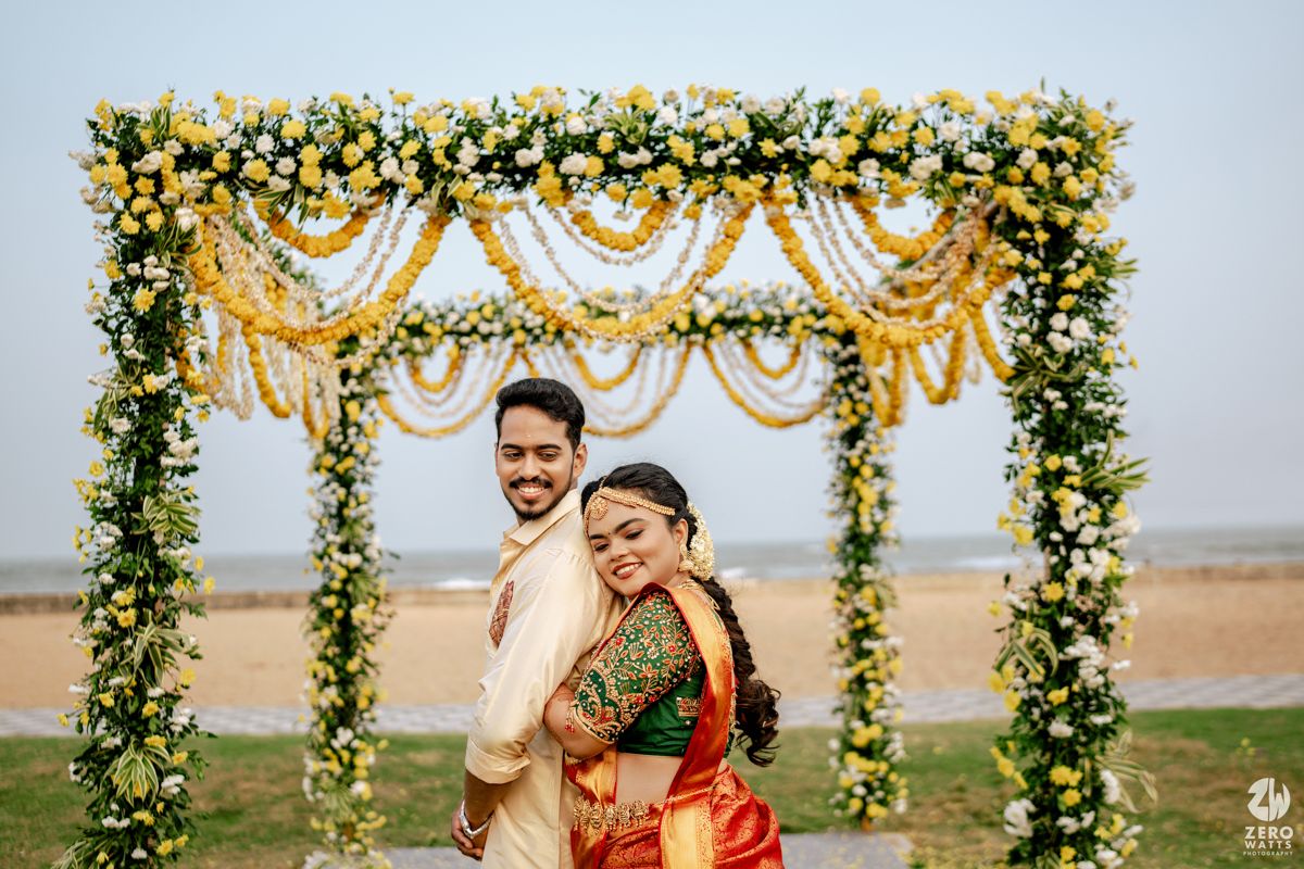 South Indian wedding couple posing together under a yellow marigold floral arch on beach.