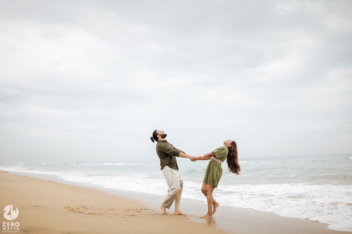 Couple holding hands and dancing playfully on a cloudy beach with waves crashing in the background.