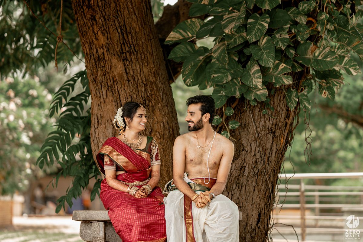 Couple in traditional South Indian wedding attire sitting together under a large tree.