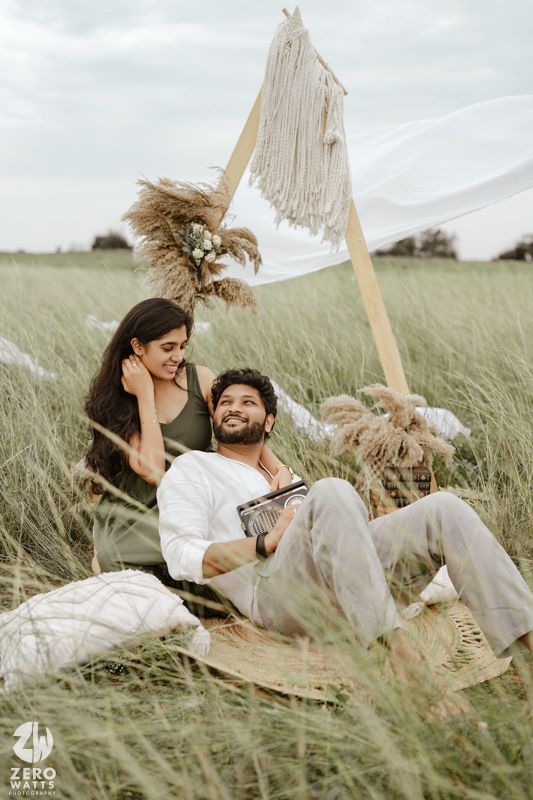A couple relaxes together in tall grass near a boho-style macrame backdrop during an outdoor photoshoot.