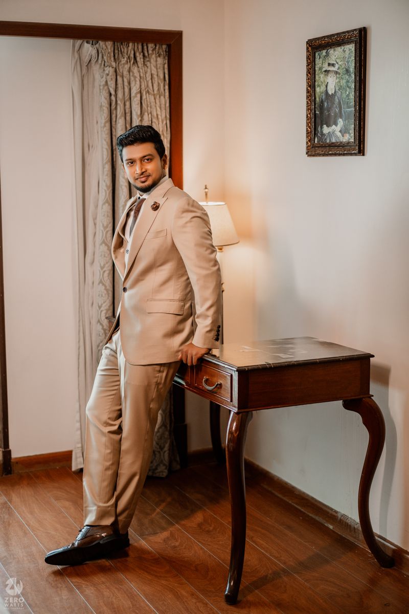 A person wearing a beige suit stands next to an antique wooden side table in a warmly lit room with curtains and artwork.