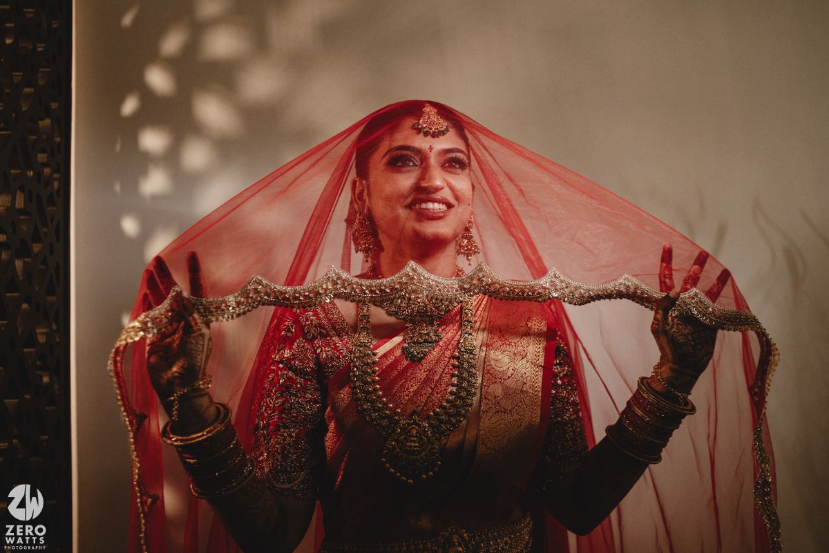 A bride in a traditional red Indian wedding dress and veil poses elegantly against a neutral background.