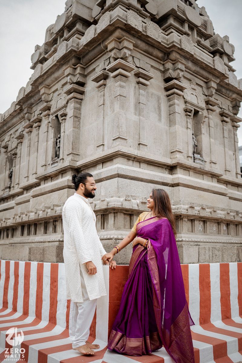 Couple in white and purple traditional attire pose against ornate temple architecture.